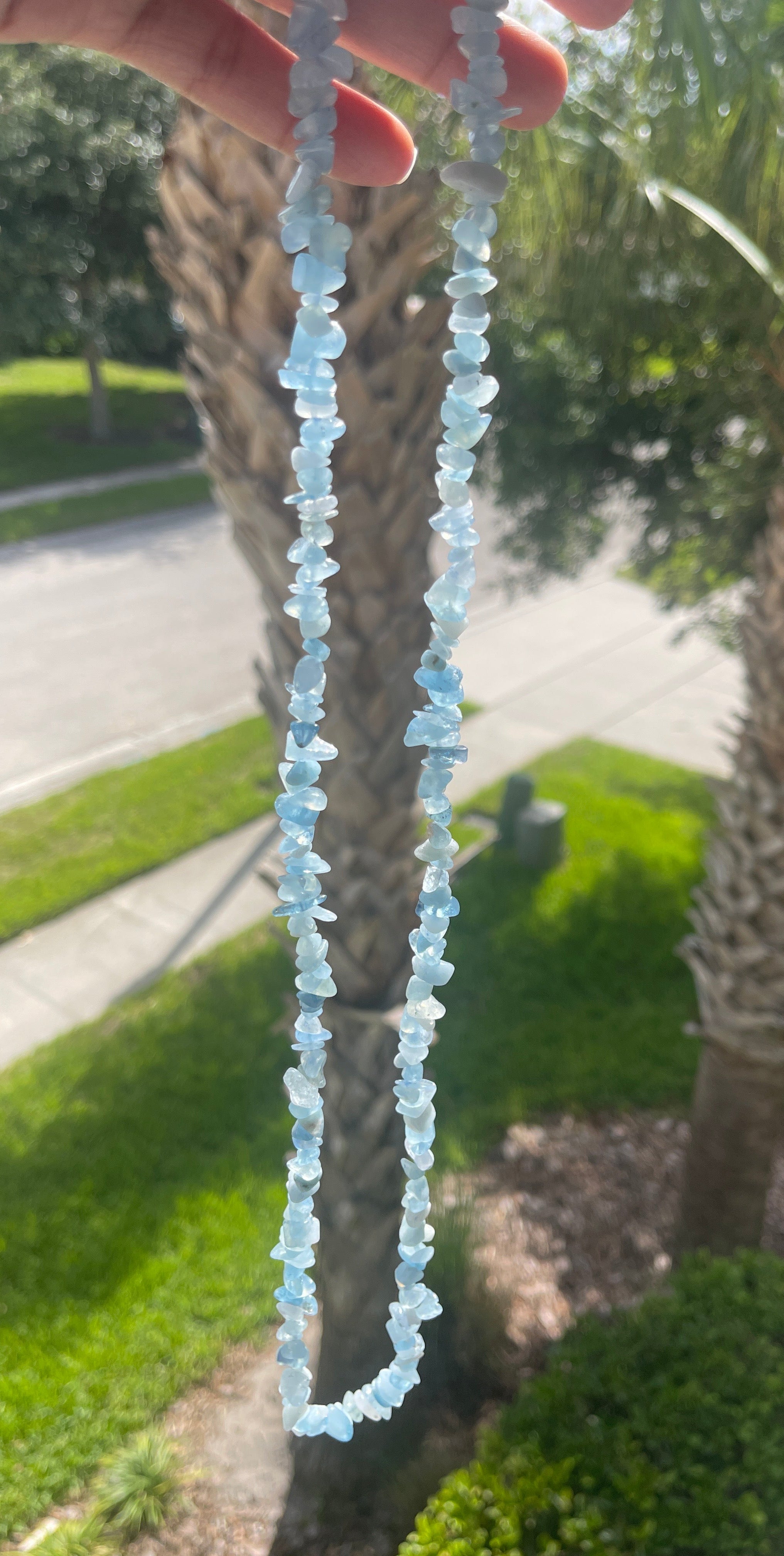 Hand holding a Aquamarine Necklace of clear and baby blue beads with a palm tree and road in the background