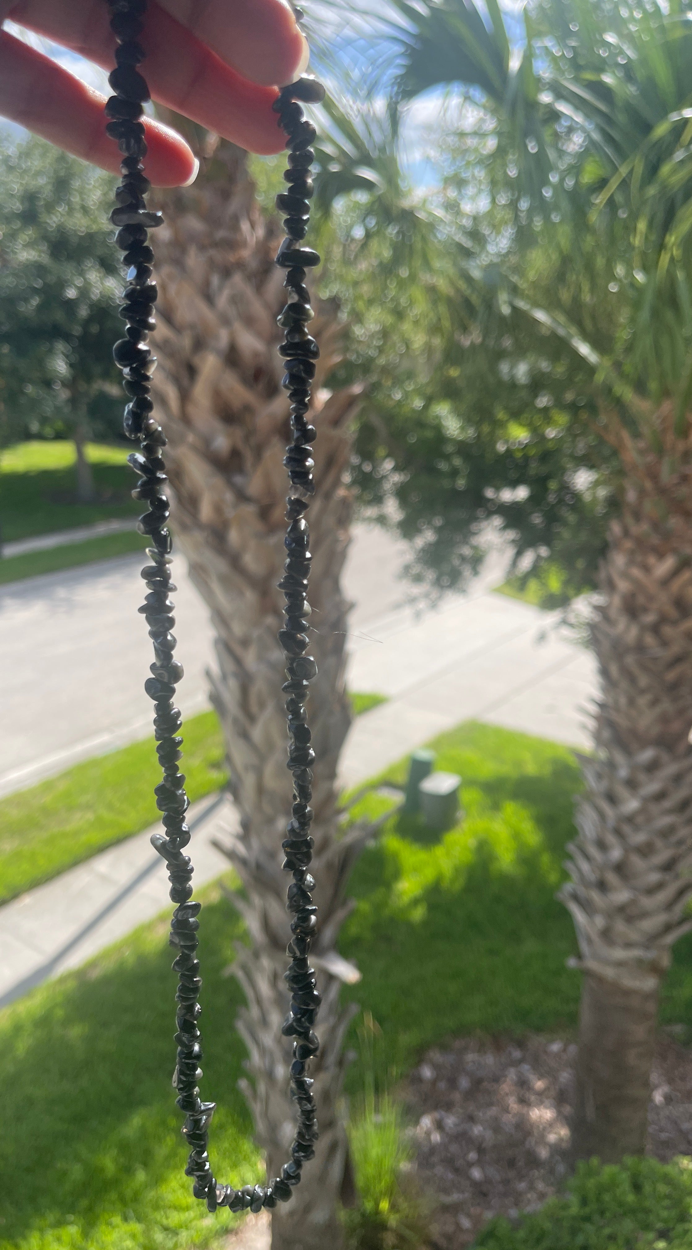 Black beaded Obsidian Chip Necklace held up to palm trees with a blurred road and greenery in the background