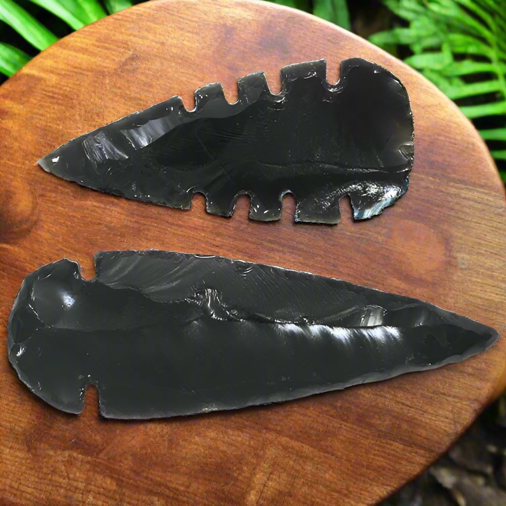 Black Obsidian Plain and Serrated Edge Arrowheads resting on a walnut table in a tropical jungle, with dappled sunlight filtering through lush, vibrant greenery.