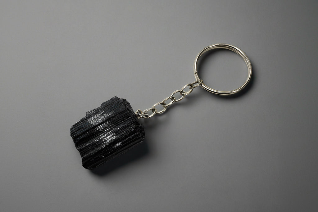 A natural black tourmaline chunk attached to a silver-colored keychain base, set against a gray background.