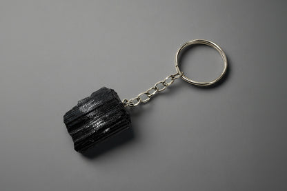A natural black tourmaline chunk attached to a silver-colored keychain base, set against a gray background.