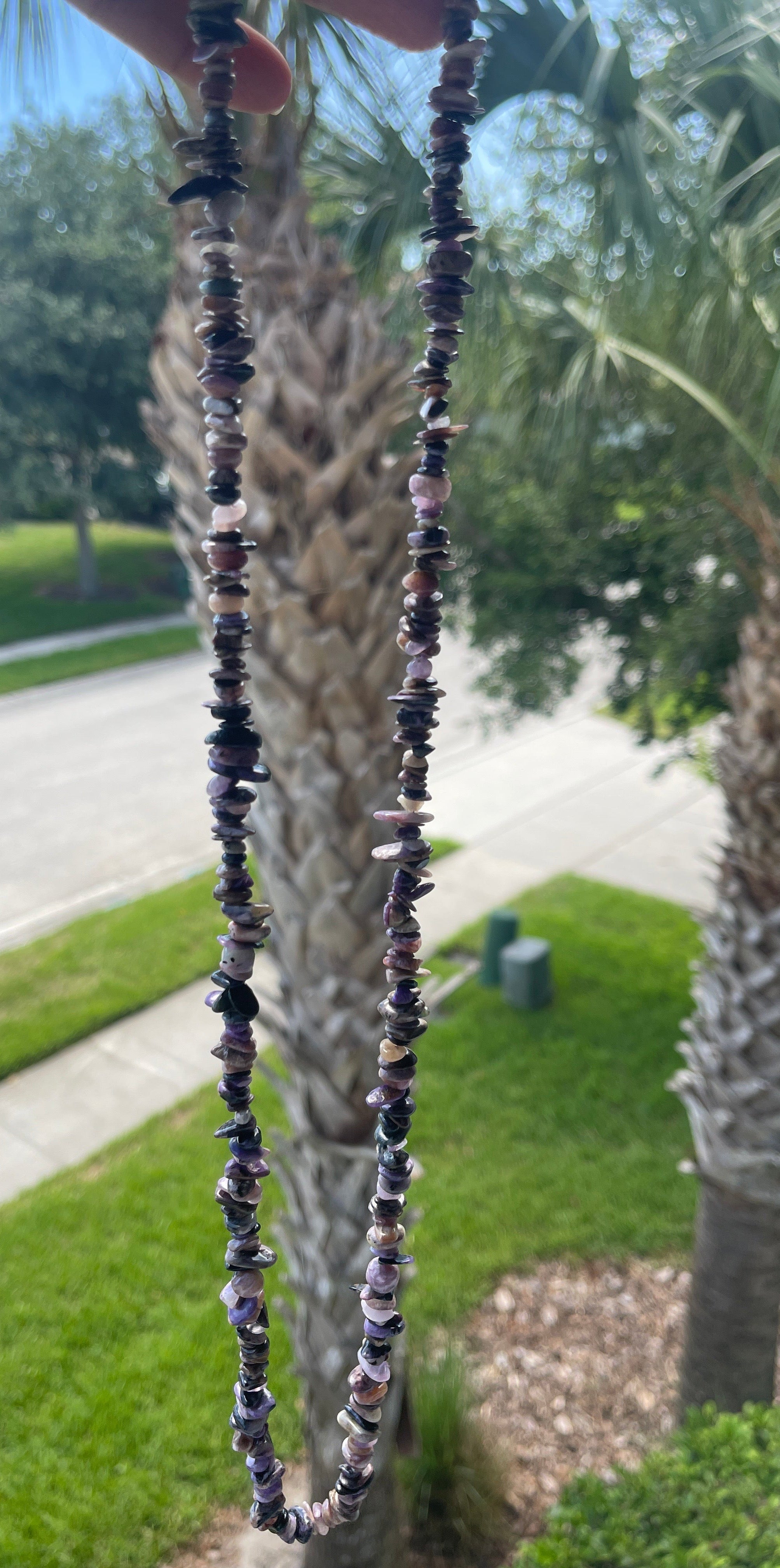 Close-up of a beaded Charoite_Necklace_30-32_Inches held against a palm tree background