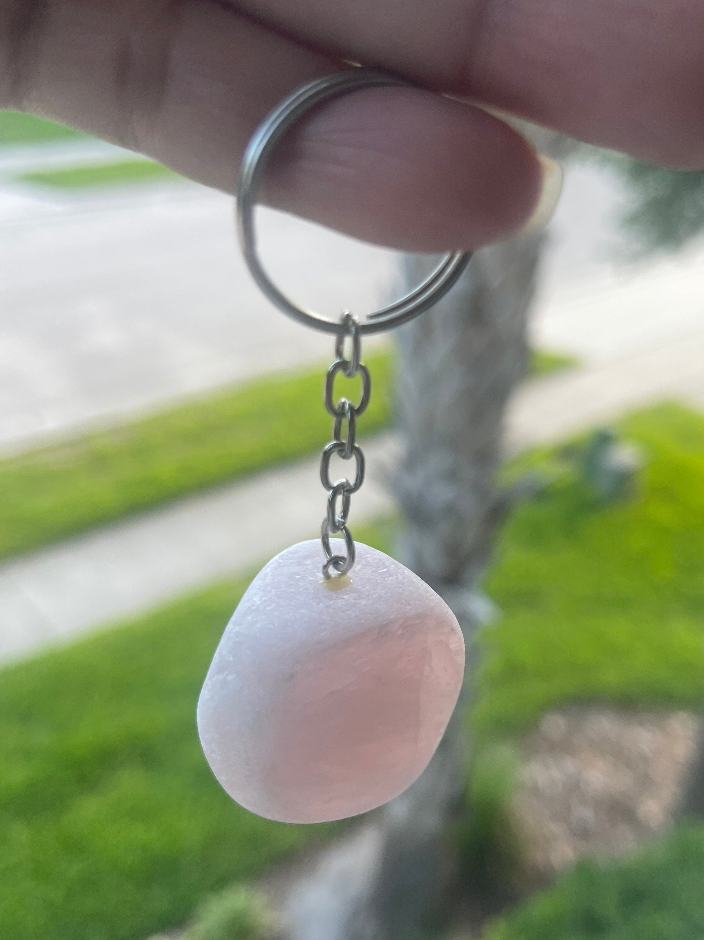 Hand holding a Ema Egg Seer Stone Polished Keychain with a pink bead against a blurred natural background
