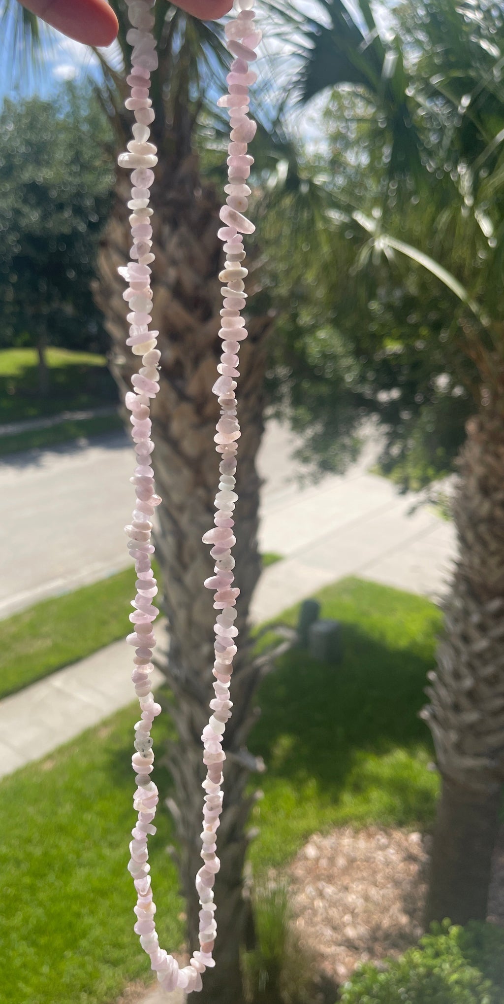 Close-up of a Kunzite_Chip_Bead_Necklaces_30-32_Inches beaded necklace held against a blurred outdoor background with palm trees.