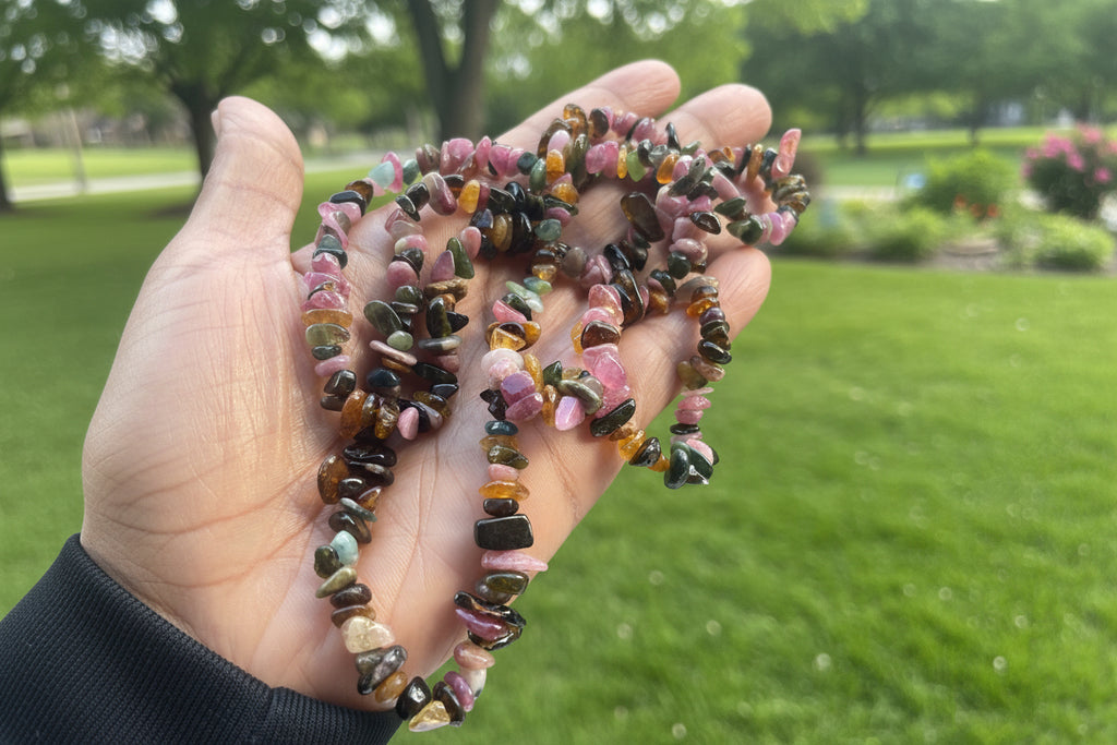 Hand holding a multicolored stone Mixed_Color_Tourmaline_Necklace_30-32_Inches with a blurred park background