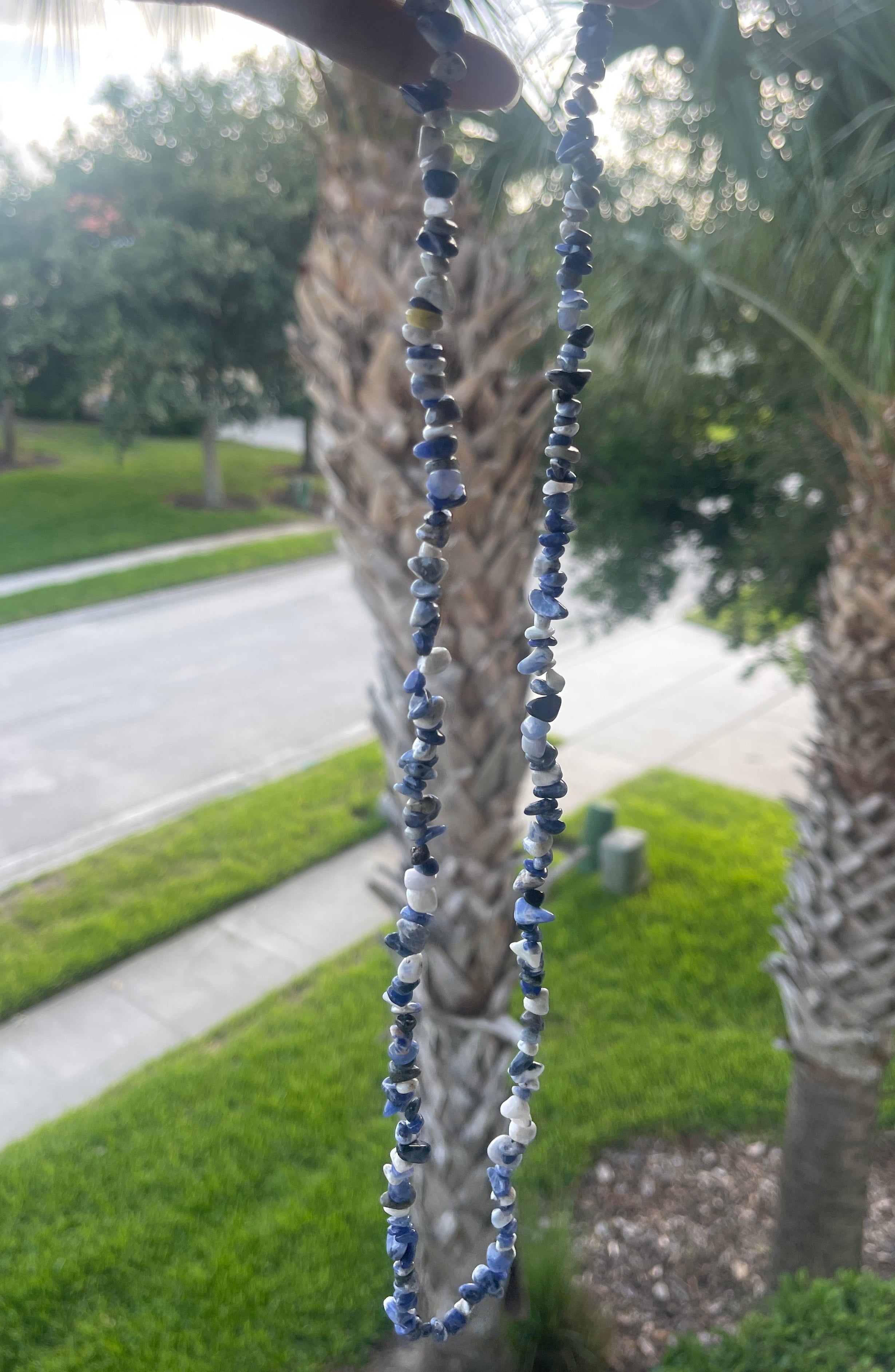 Hand holding a string of blue beads Natural_sodalite_chip_necklace  with palm trees and a road in the background