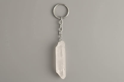 A clear quartz crystal point attached to a metal keychain ring, with the crystal hanging vertically against a gray background.