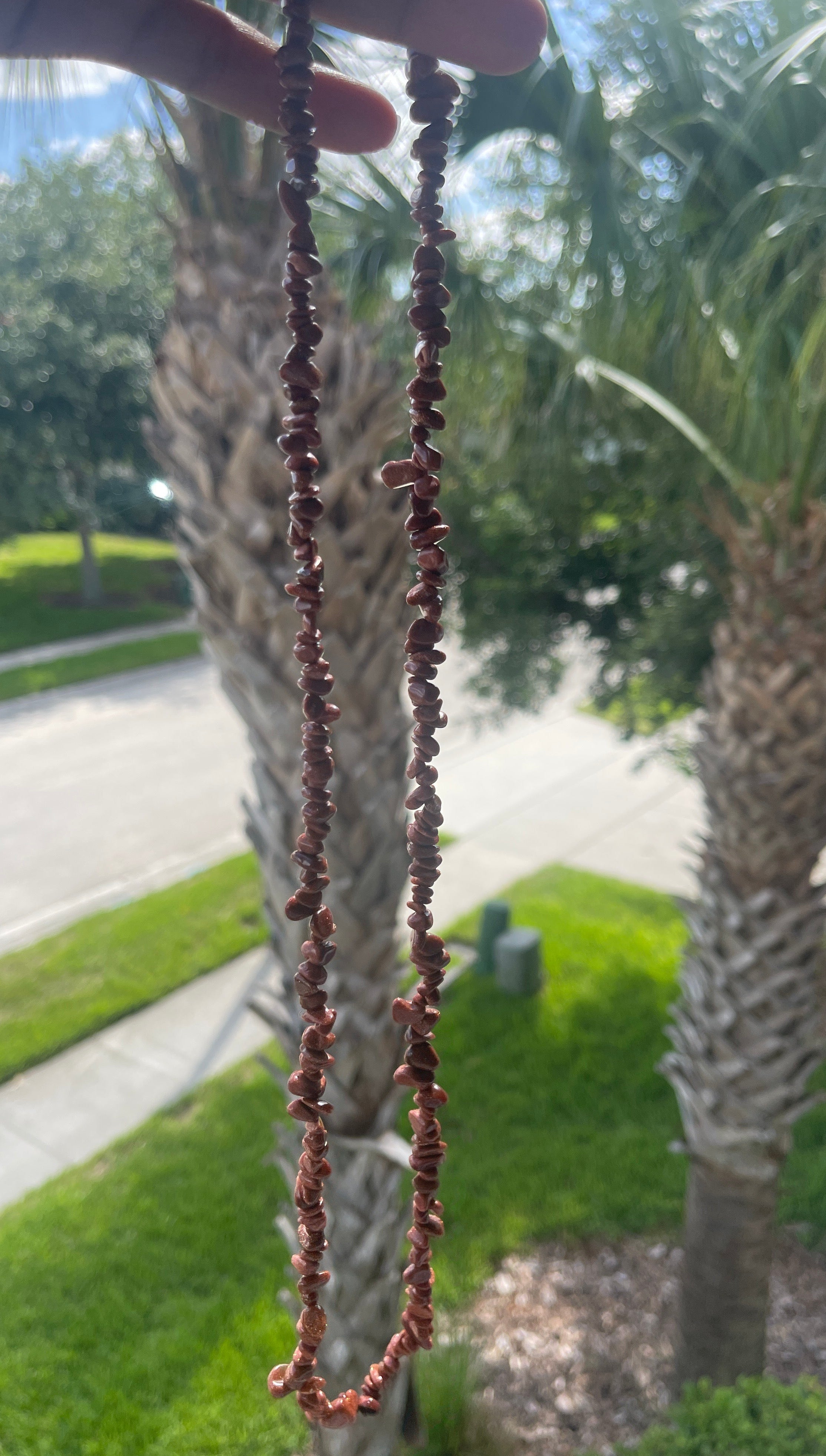 Person holding a Red_Goldstone_Necklace_30-32_Inches of beads with palm trees and a road in the background