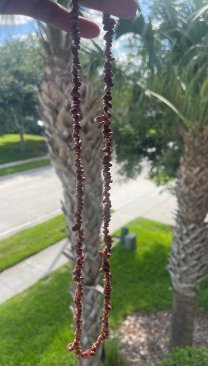 Person holding a Red_Goldstone_Necklace_30-32_Inches of beads with palm trees and a road in the background