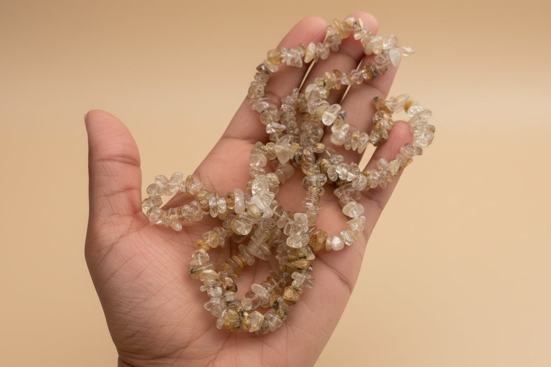 Hand holding a Rutilated_Quartz_Necklace_30-32_Inches with a blurred tan  background