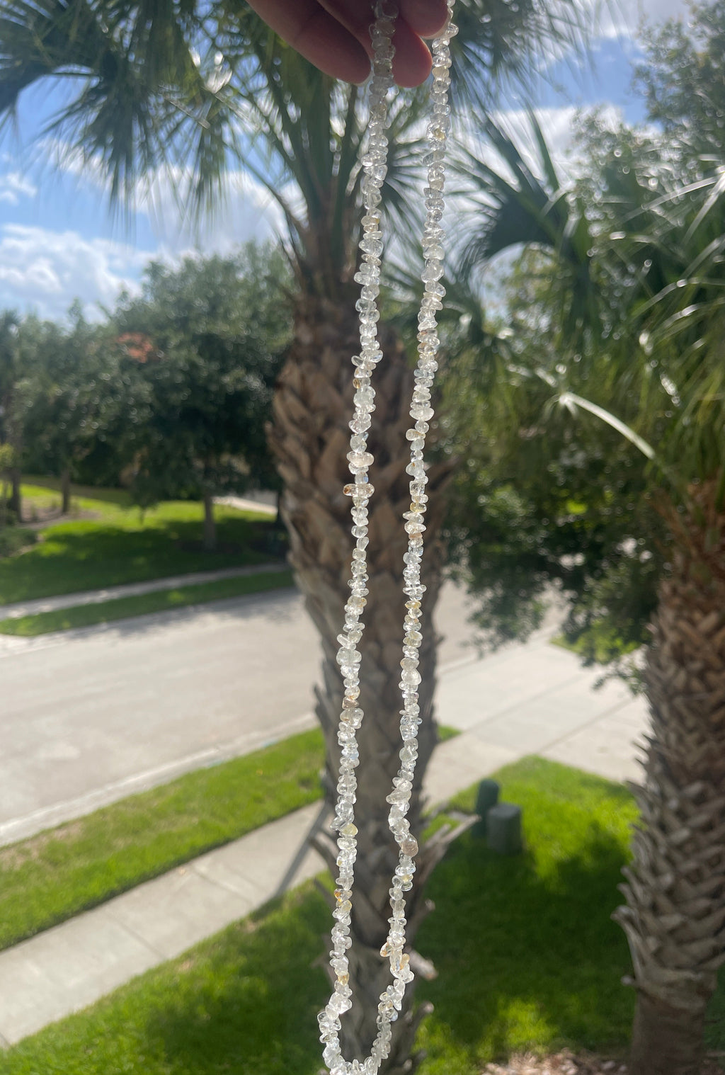 Person holding a Topaz string necklace in sunlight with palm trees and a road in the background